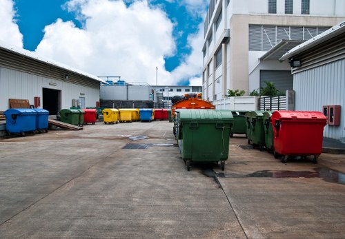 A broad view of an industrial or commercial waste collection area featuring multiple wheeled bins in various colours including green, red, blue, yellow, and grey, arranged along a paved surface with visible expansion joints. The bins are positioned in front of a white warehouse building with a flat roof and metal siding, and are situated on either side of an orange waste container or vehicle part. The background includes another industrial building with a façade of large windows and horizontal louvers, under a partly cloudy sky with patches of blue. The area appears clean and organized, reflecting a professional rubbish removal service environment typical of commercial waste management operations, with the external setting possibly within an urban or suburban context in the Balham region. This scene aligns with waste collection activities managed by Commercial Waste Removal Balham, offering insight into the typical layout used for rubbish disposal in this locality.