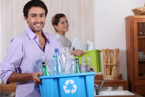 A young man with dark hair, a beard, and wearing a light purple shirt is smiling and holding a blue recycling bin filled with mostly glass bottles, including clear and green ones, in a domestic kitchen environment. Behind him, a woman with brown hair tied back, wearing a light-colored top, is standing near a green plastic box, with a wooden cupboard and shelves visible in the background. The kitchen has a neutral decor with wooden furnishings and a beige wall, and the scene is well-lit with natural or artificial light. The blue bin features a white recycling symbol. This image relates to rubbish collection and waste disposal services focused on recyclable materials, indicating a household effort to separate waste for proper disposal, characteristic of professional waste removal providers operating in residential areas.