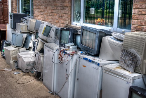 The image displays an outdoor disposal area with a long row of old, discarded computer monitors, CRTs, and electronic waste piled against a brick wall adjacent to a large window. The monitors vary in size and shape, with many showing signs of wear such as yellowed screens, rusted or frayed cables, and dust accumulation. Some monitors are stacked on top of each other while others lean against the wall, creating a cluttered appearance. The concrete ground beneath the waste is visible, with a few loose cables and electronic components scattered around the bases of the devices. The background features the brick exterior of a building and the reflective glass of the window, which shows hints of greenery outside. The scene illustrates electronic waste that requires professional rubbish removal, similar to services provided by Commercial Waste Removal Balham, in a setting that reflects the need for proper disposal of outdated electronics to ensure environmental compliance and local waste management standards in Balham or nearby areas.
