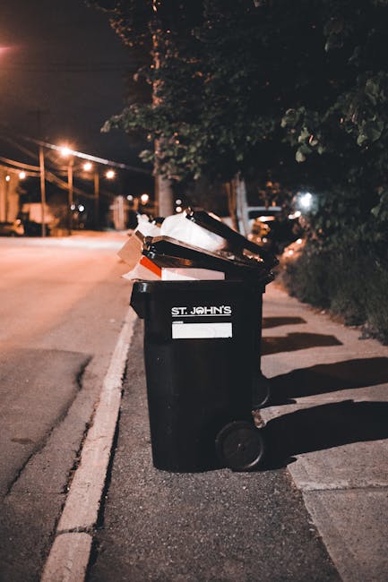 A black wheeled rubbish bin positioned on a sidewalk beside a curb in an urban environment at night, with its lid open and filled with assorted waste, including paper and cardboard. The bin is marked 